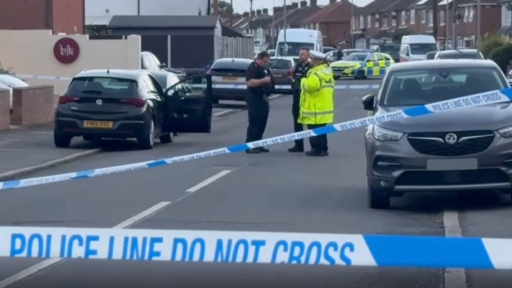 A residential street cordoned off with blue and white police tape. Three police officers, one in a hi-vis jacket, stand near a black car with its driver's door open. A police car further away is parked, blocking the road.