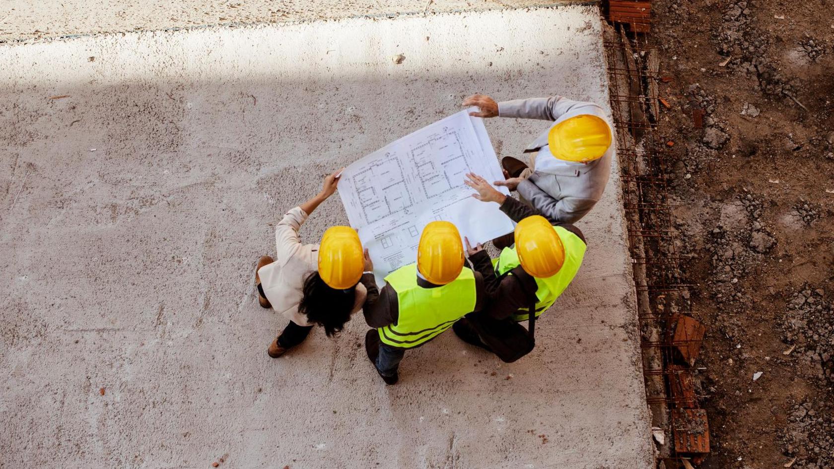 A stock image of four people on a construction site standing on a large slab of grey concrete. They are holding out a large roll of white paper which displays plans of a house on it. They are wearing yellow hard hats, and two of the people in the middle are wearing yellow hi-vis jackets.
