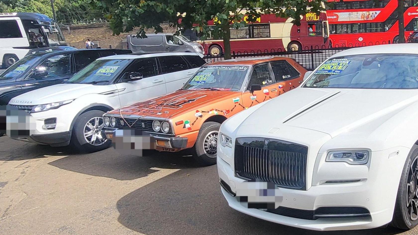 A row of seized cars with a white Rolls Royce closest on the right hand side of the picture, next to it an orange Nissan Datsun Skyline, then a white Range Rover. In the background are two buses and a silver van driving along a road.