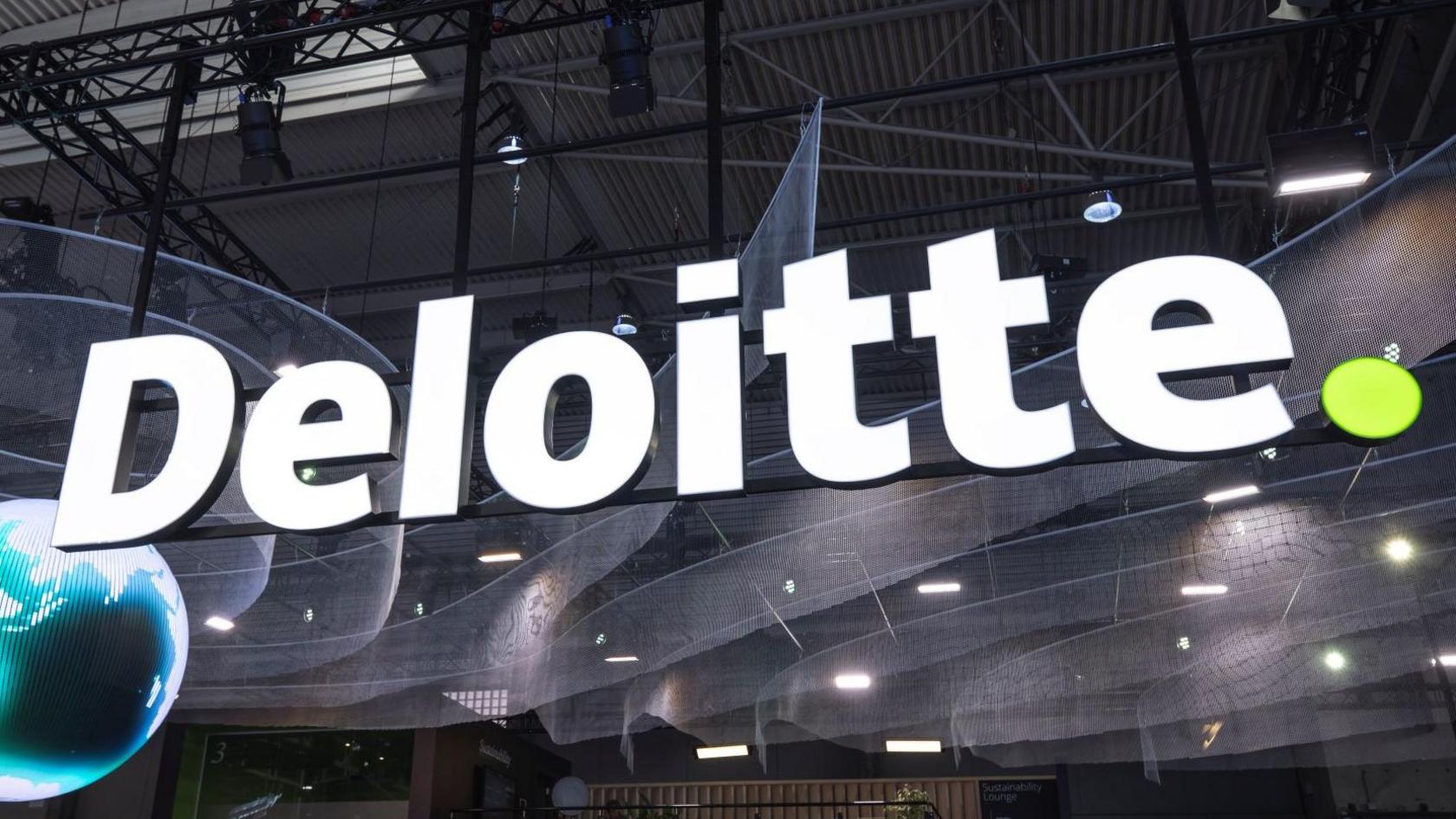 A large Deloitte sign in white letters with a small green full stop at the end. The large lighted sign is hanging over a large hall floor and there is a pixelated globe on the left of the picture.