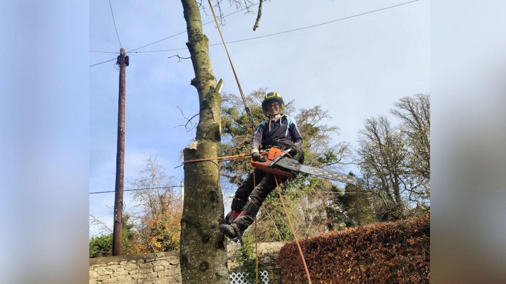 A person is in the middle of a tree. They are attached to the tree with ropes. They are also wearing protective clothing. The sky behind the tree is bright blue with white clouds. There are more trees in the background. There is 3 electric cables behind the tree. She is smiling at the camera.