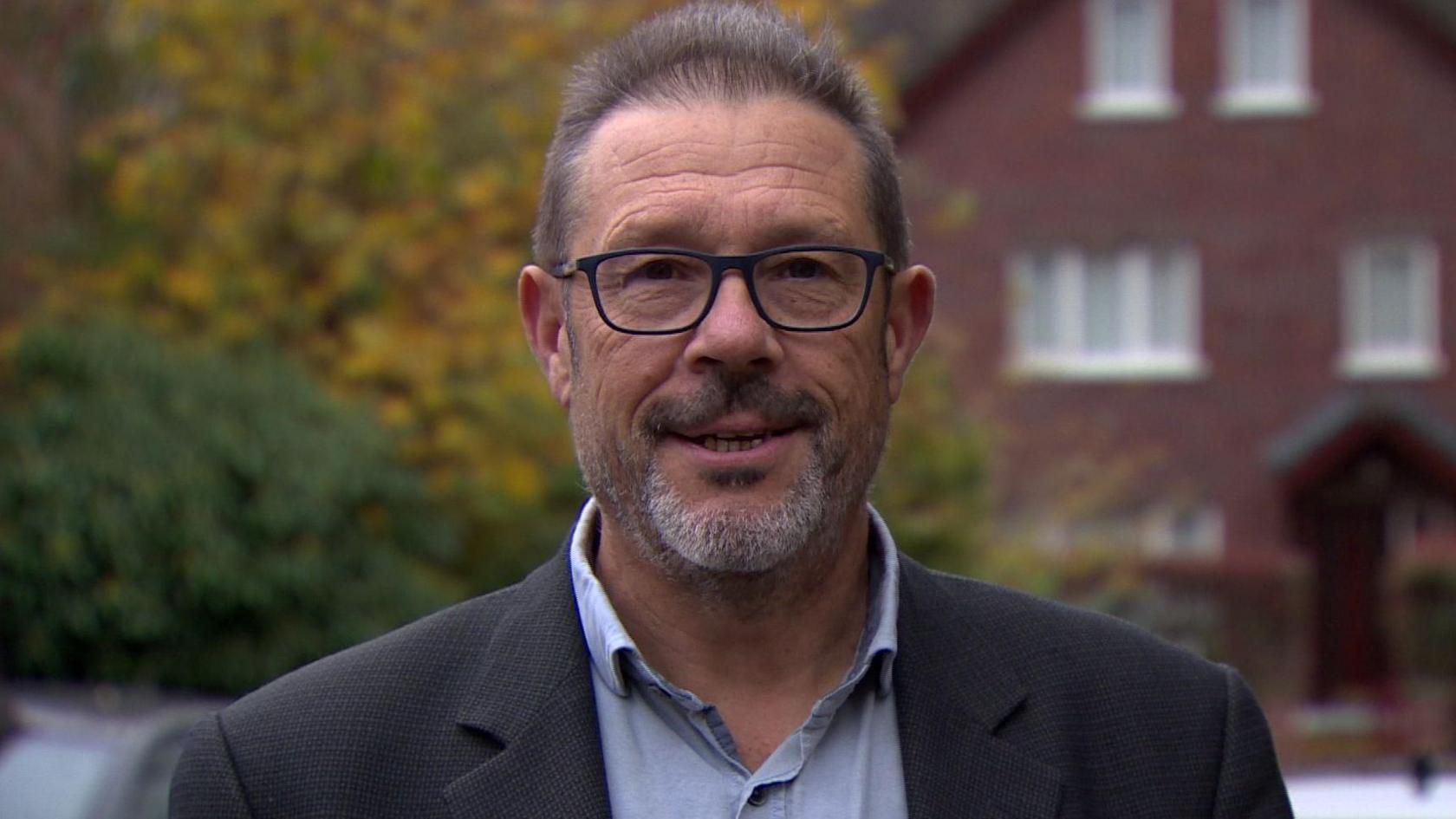 Prof Dominic Bryan, who was co-chair of Stormont's flags commission, wearing a dark blazer and blue shirt and glasses, who is pictured standing with bushes and red-brick housing in the background