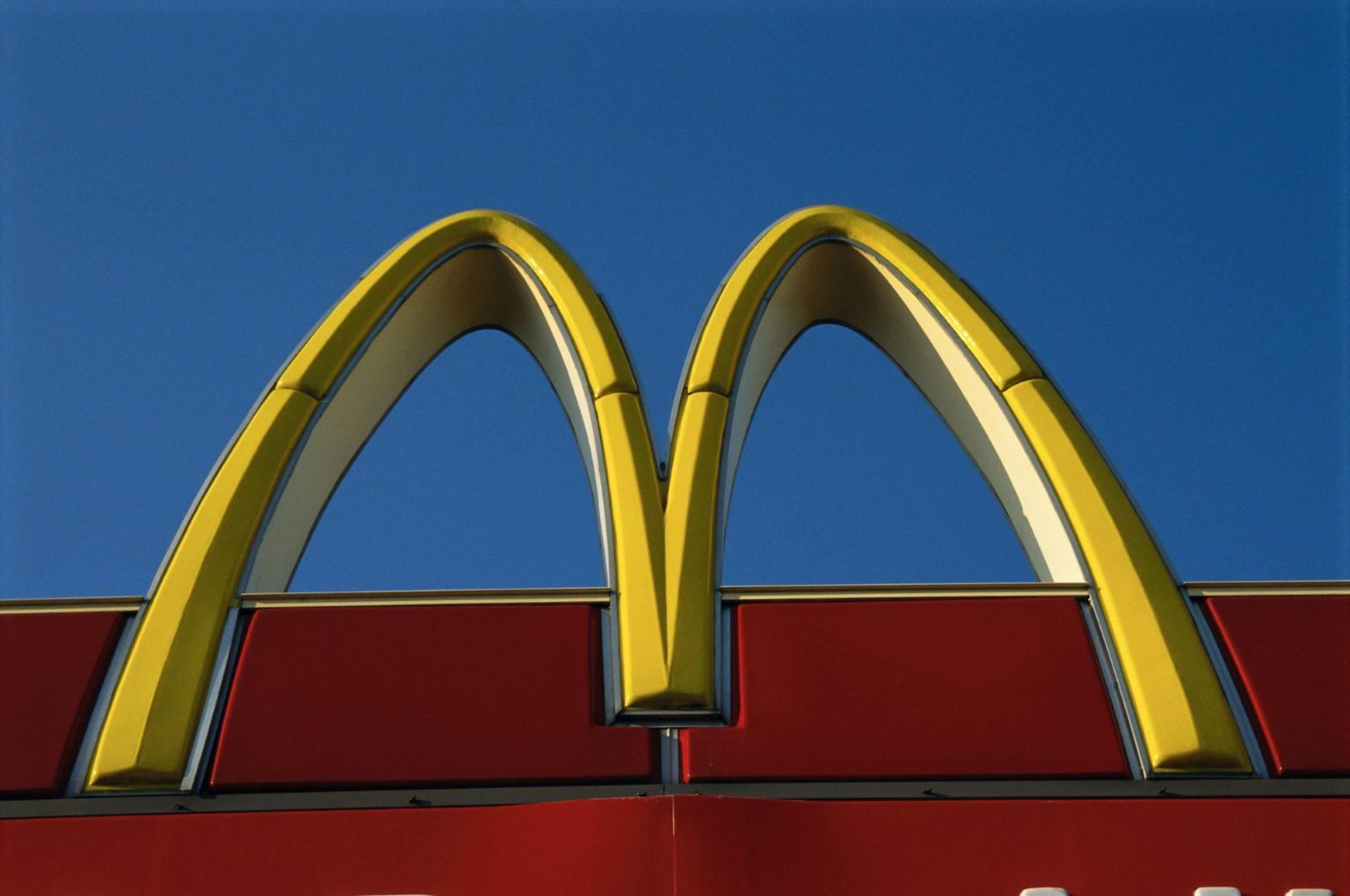 A close-up photograph of a yellow "M" from a McDonald's sign. The yellow M is on a red storefront and is against a blue sky.