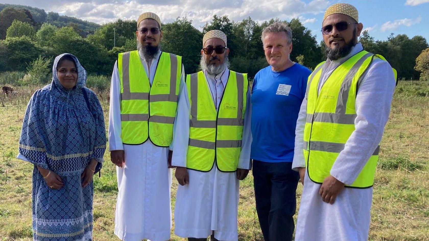 A woman wearing a blue headscarf and outfit alongside four men men wearing florescent vests and one wearing a blue sweatshirt all standing in a grassy field.