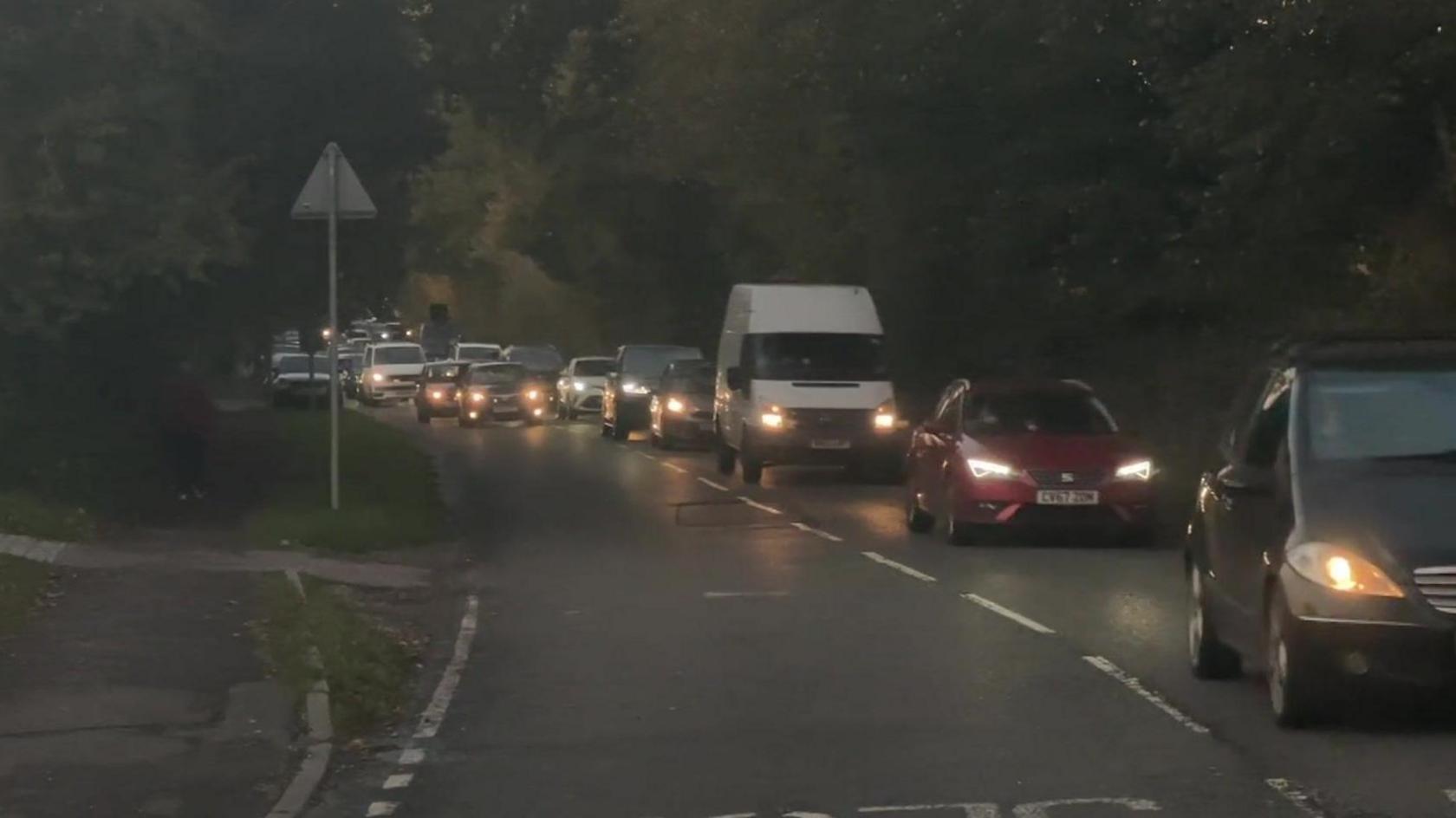 A road at dusk with cars queuing along one lane and others further back seen trying to pass them. The cars have their headlamps on and the road has thick trees on either side.