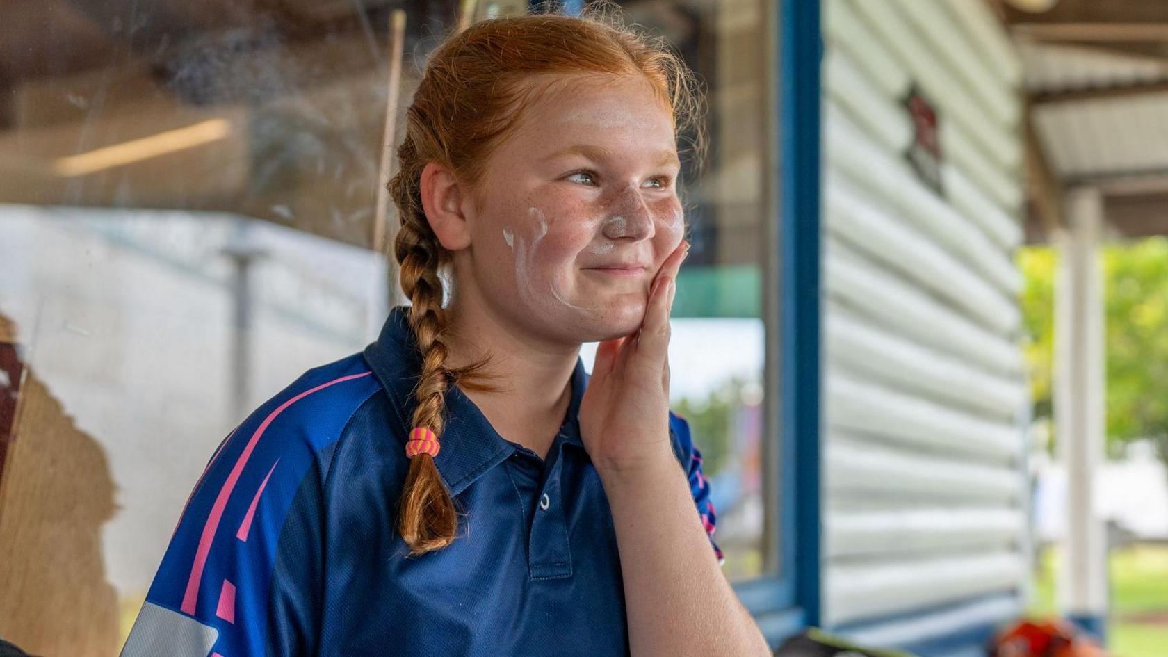 A young cricket player, her red hair braided over her shoulder, rubbing in sunscreen