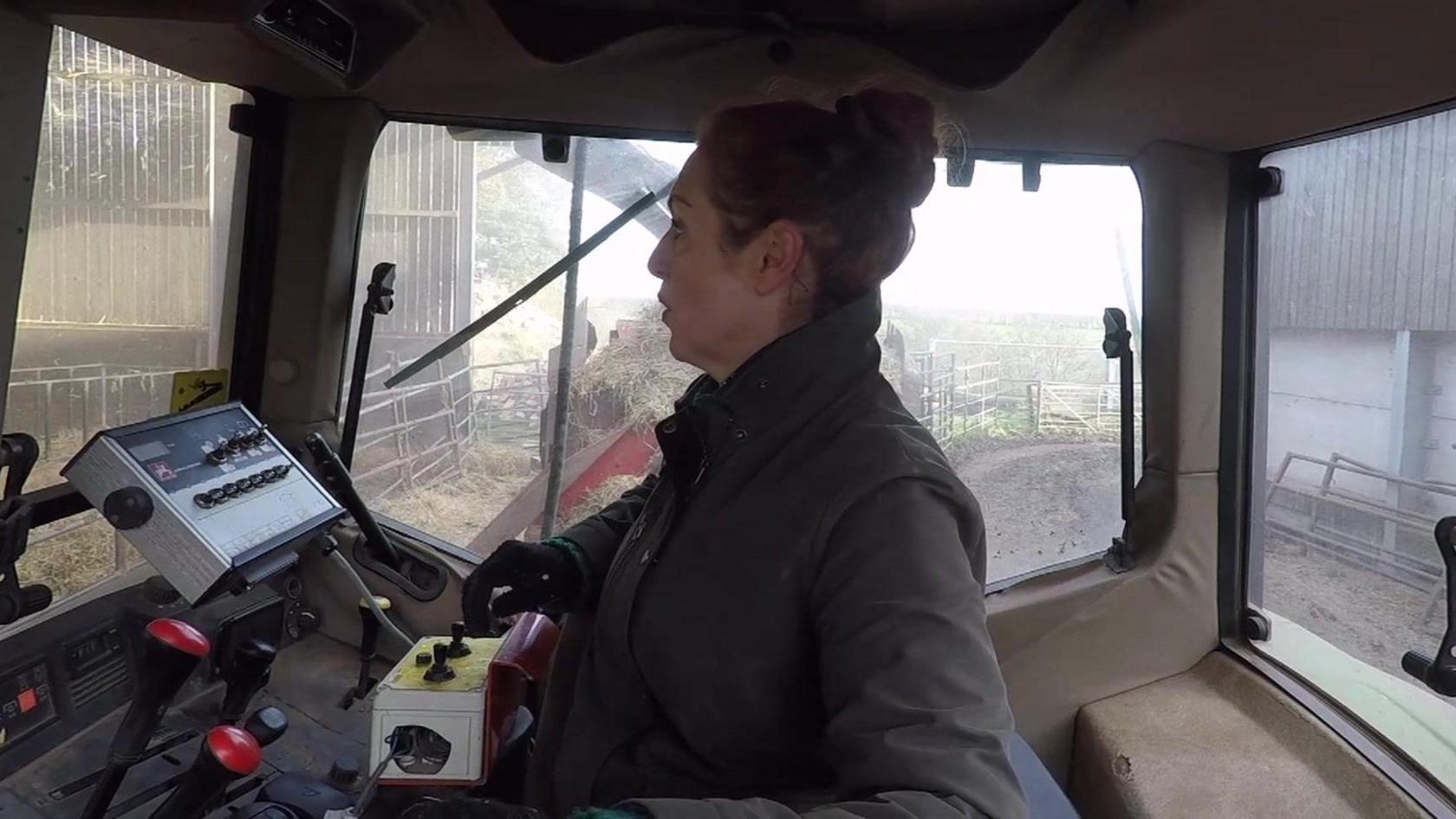 Clare Wise is sitting in the cab of a tractor, which is parked in a farmyard. 