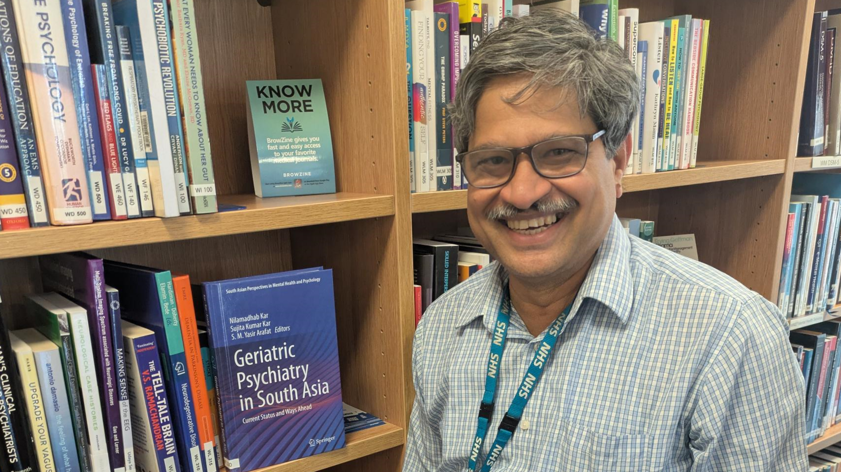 A man with grey hair, a grey moustache and glasses, wearing a blue and white checked shirt and a blue NHS lanyard, is standing next to a bookshelf containing psychiatry and psychology books, including a purple one reading "Geriatric Psychology in South Asia".