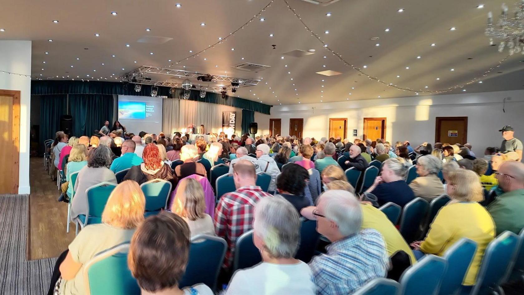 A large group of people say in a room while a meeting takes place. Guests are sat on blue chairs and a projection screen has been set up at the front of the room with four people on the stage.