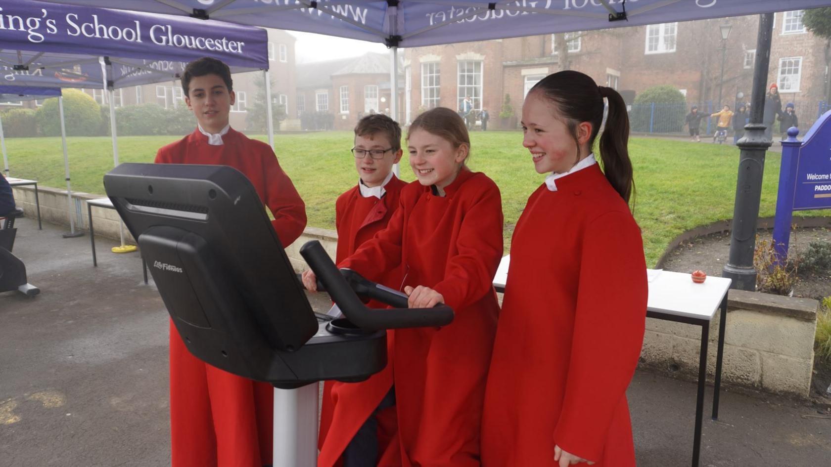 A group of four members of the school choir, dressed in bright red robes with white collars. One of the girls is sitting on the static bike and looking at the electronic screen, while the others stand beside her smiling at encouraging her. The weather is grey, wet and misty. The bike is positioned underneath a blue cabana which says The King's School Gloucester on it.