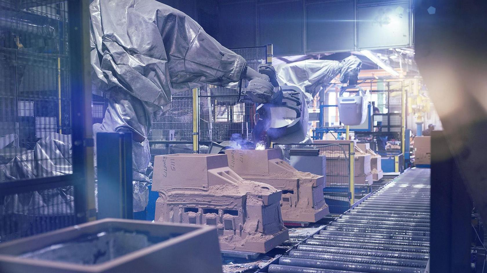 A room with heavy machinery and sand casts. A conveyor belt made of metal rods is on the right with several large industrial machine to one side with a wire fence behind them.