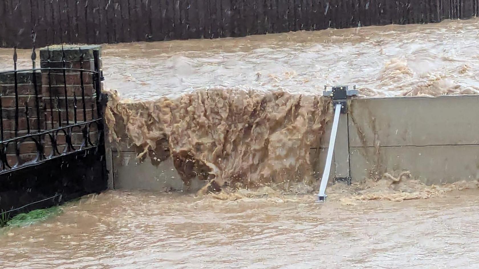 Flood water goes over the top of a flood barrier outside a house in Chard in Somerset. The flood water is a light brown colour and the barrier is grey. A stone wall and dark railings are also visible