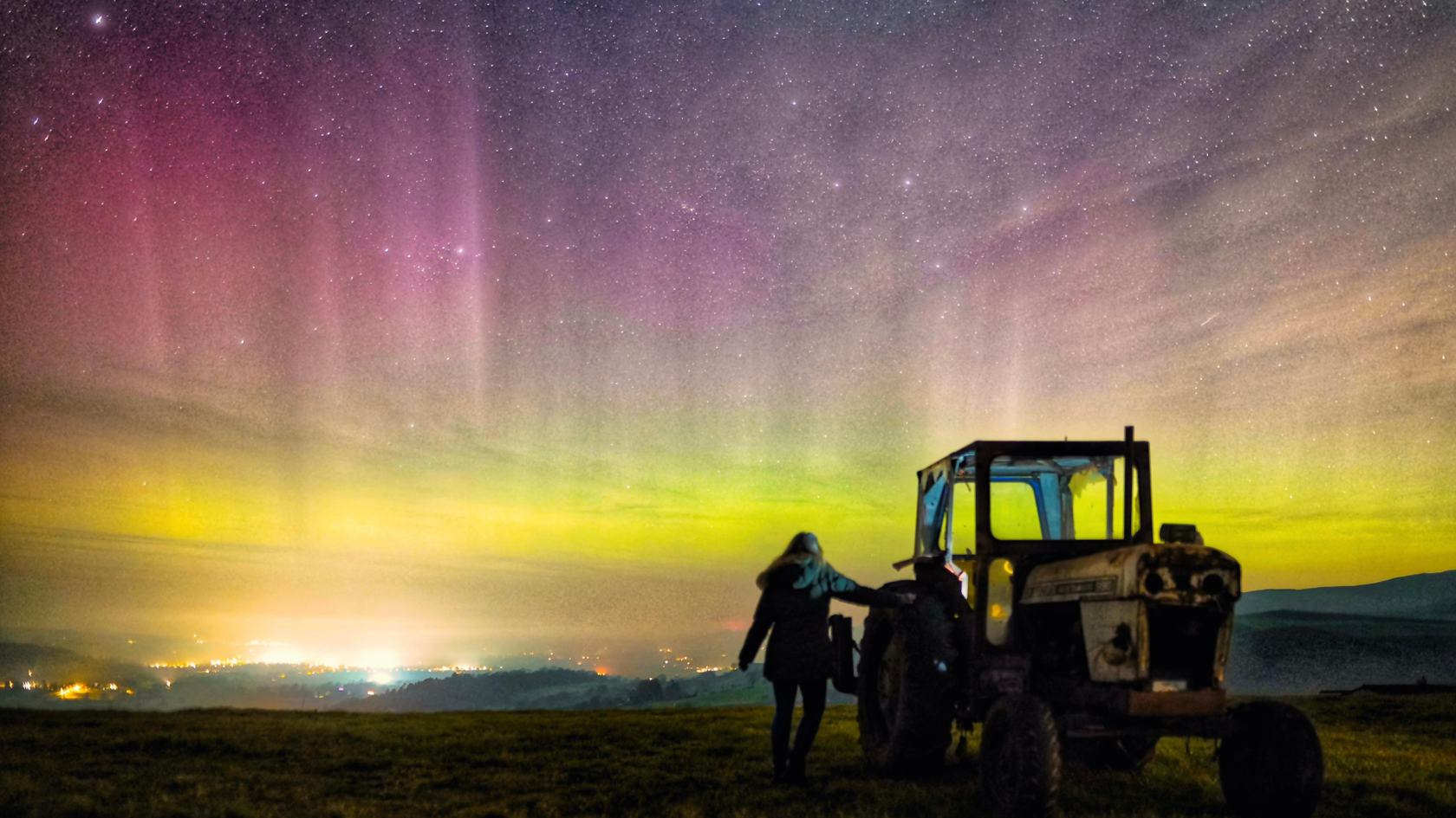 person standing next to a tractor with the bright lights of the northern lights in the night sky. Green and purple streaks fill the sky
