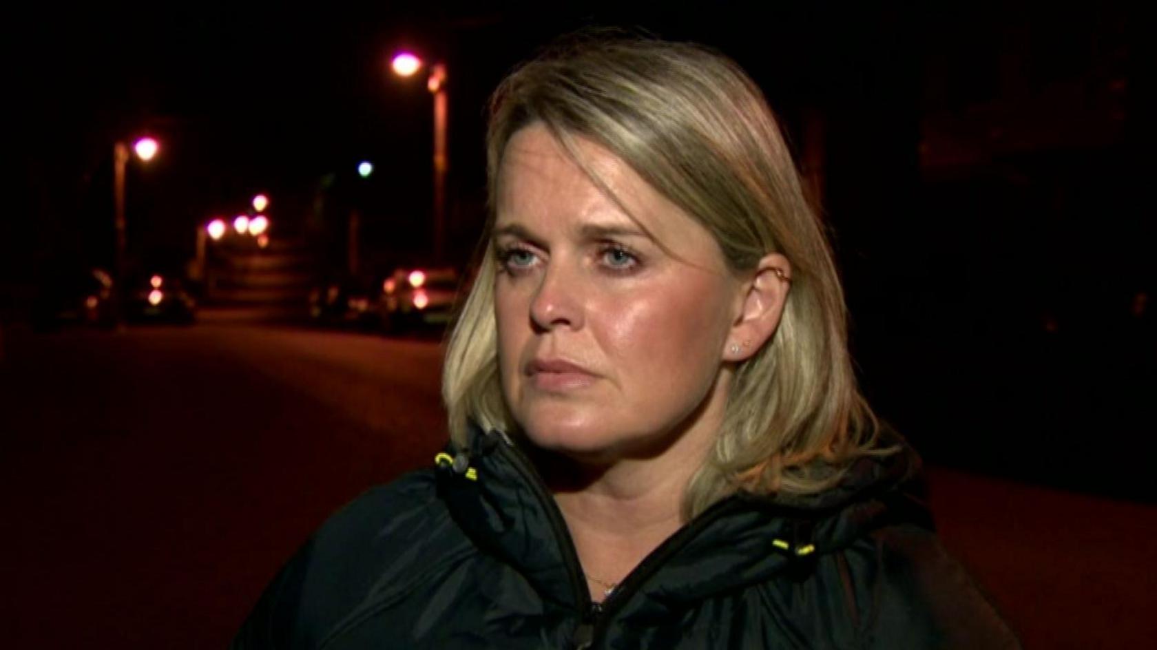 Clare Loughran speaking to a BBC reporter on a dimly-lit street. She has shoulder-length blonde hair and is wearing a dark-coloured raincoat.