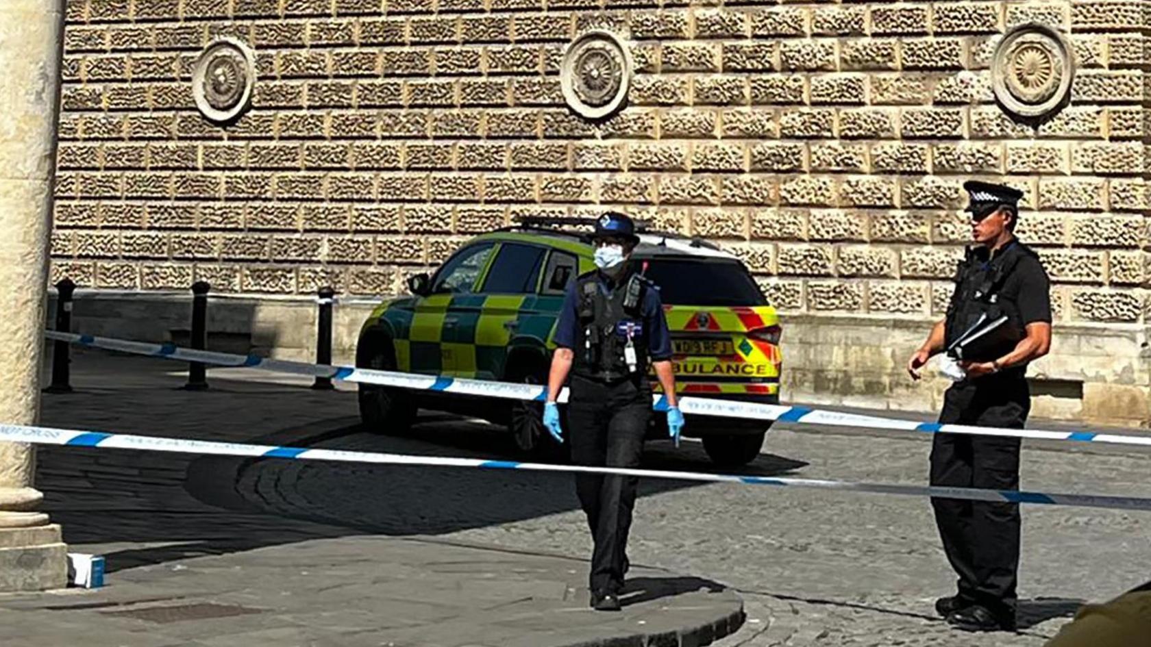 Two police officers in Bath. Police tape can be seen, blocking access to the public. One officer is wearing a face mask and blue gloves. The other is standing nearby, with no PPE. An ambulance car can be seen behind them both. 