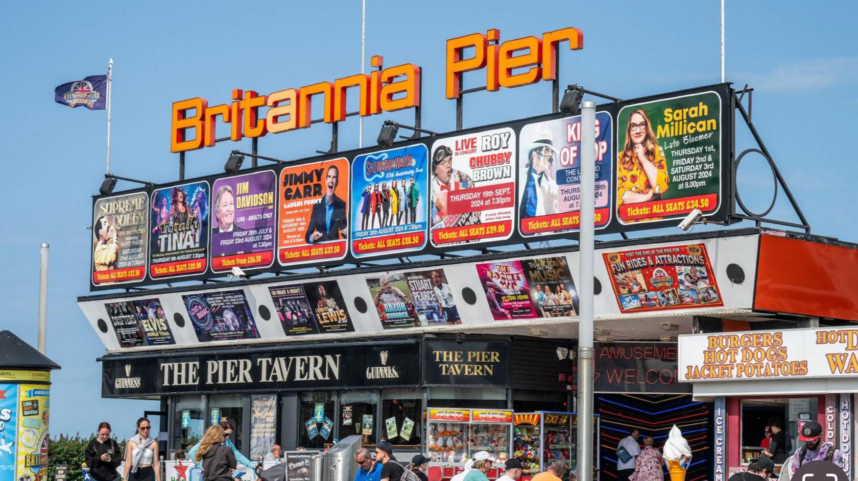 View of Britannia Pier from the front on the right. A orange sign is above the entrance that says Britannia Pier, and multiple posters that show who is set to be at the pier.