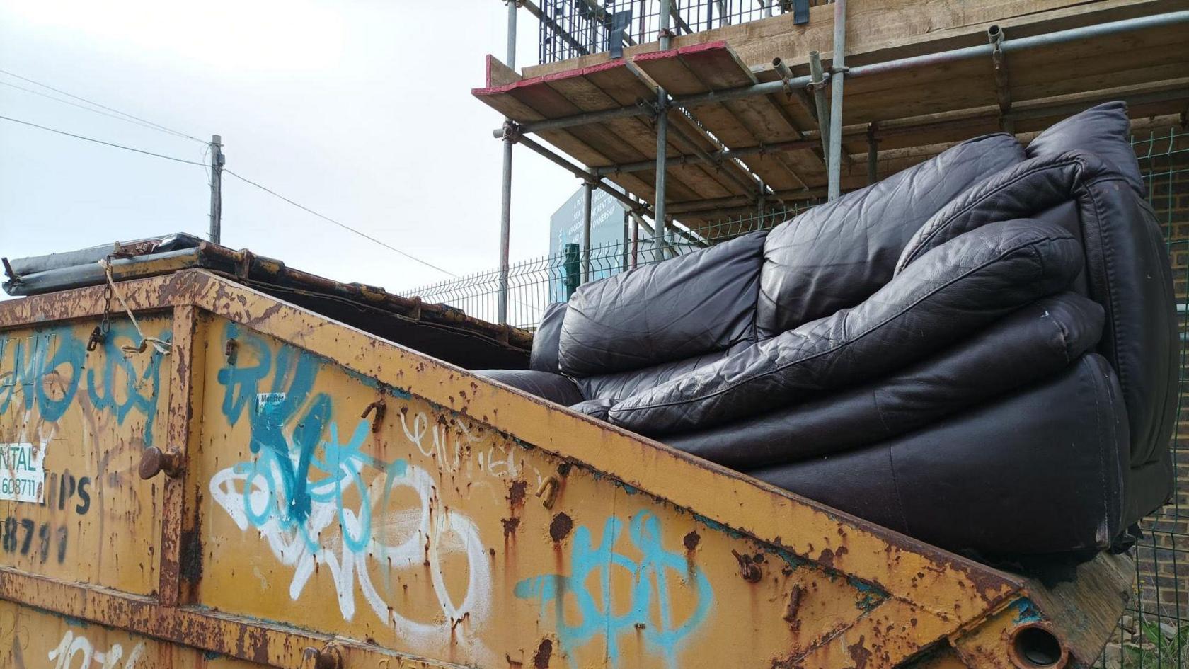 A sofa hangs out a yellow skip with graffiti. 