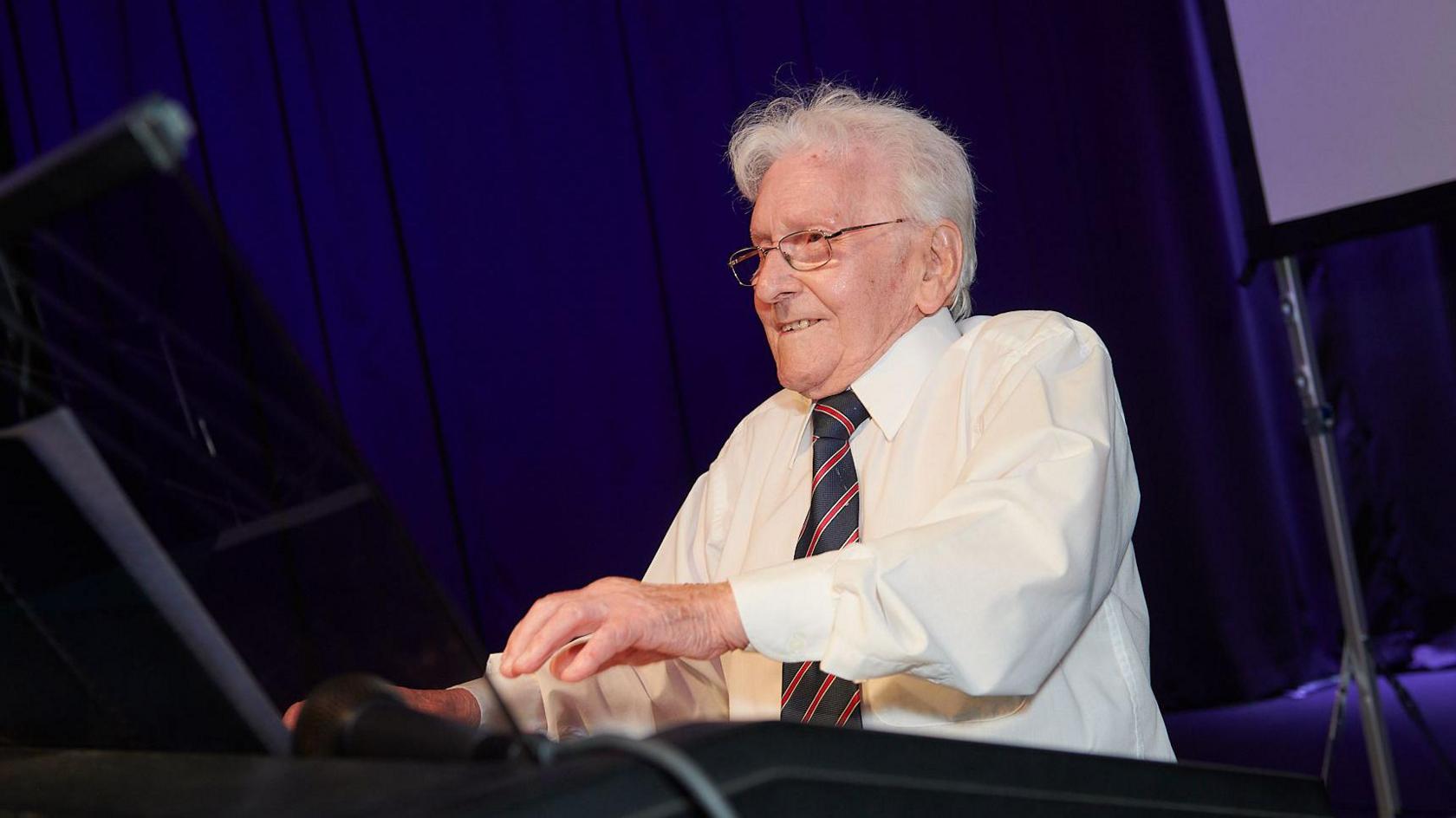 An elderly man with white hair, wearing a white shirt and striped blue and red tie, sits with his hands out, ready to play an instrument