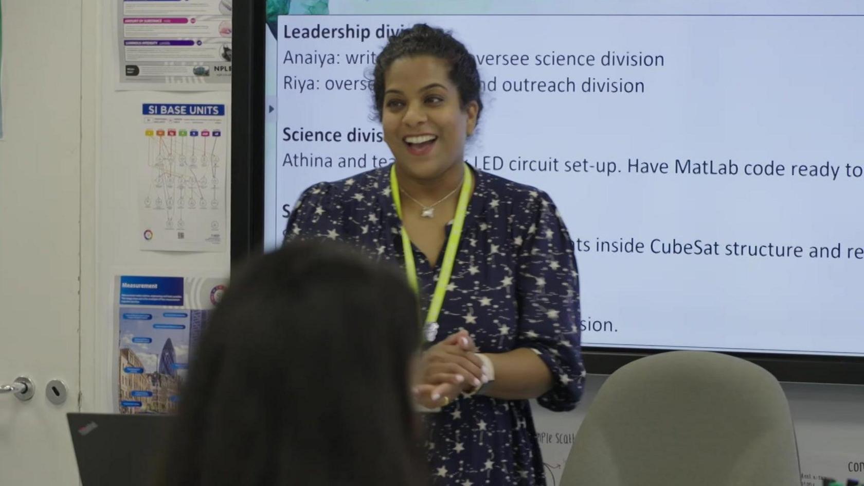 A teacher standing at the front of a classroom in front of a whiteboard. She is wearing a blue patterned dress and a yellow lanyard. The back of the heads of some pupils are in the foreground. 