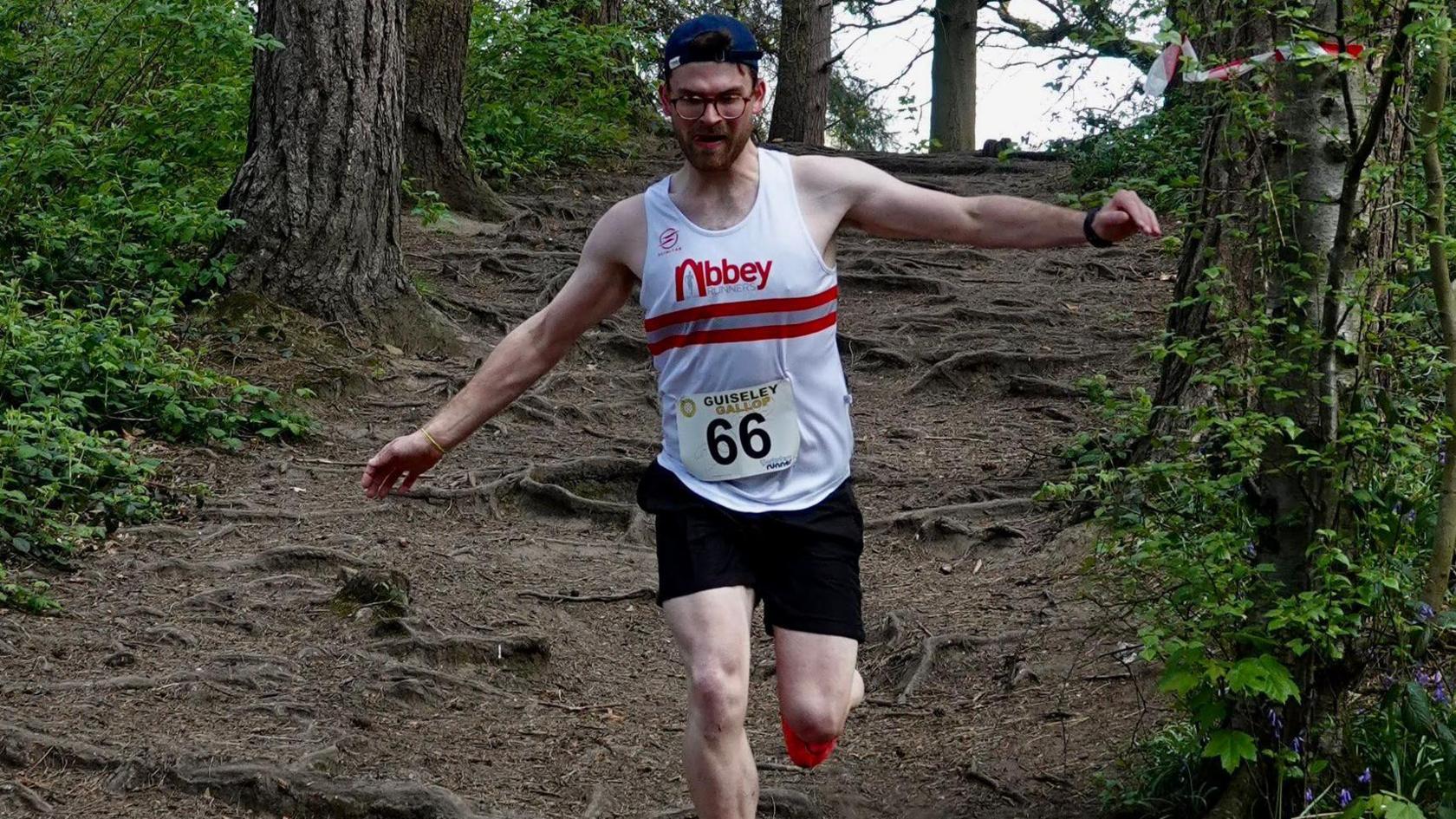 Euan Holwill running through woodland. He is wearing a backwards blue cap and a white vest which reads 'Abbey runners'.
