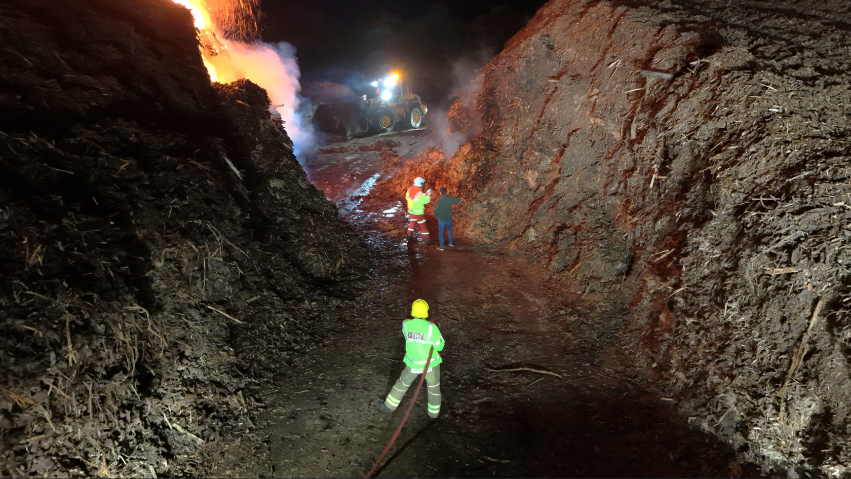 A firefighter stands between two large piles of bark which are much taller than him, with a hose under his right arm. The image is taken from behind him, with two men and a digger further in front of him, with the pile on the left on fire.