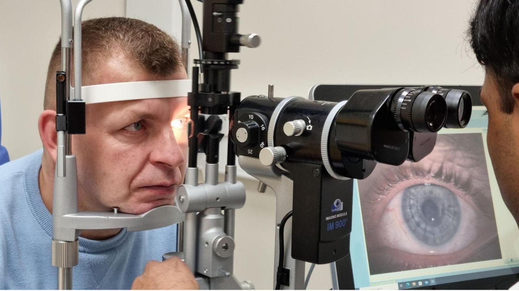 Tomasz is sat in a blue chair and is wearing a light blue sweatshirt. He has blue eyes and short brown hair. He is resting his chin and forehead on a metal frame. In front of the frame, a man is using what looks like a large pair of black binoculars to scan Tomasz's eyes. This brings up a close-up image of his eye on a computer screen. 