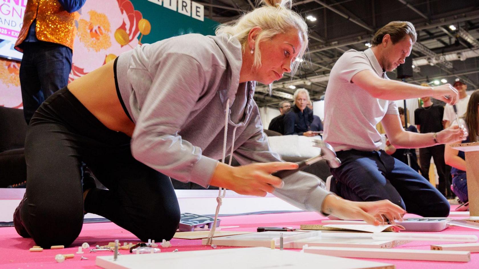Hayley McAuley, who has blonde hair, a grey hoodie and black jogging pants is kneeling down and holding a hammer as she assembles a piece of flatpack furniture