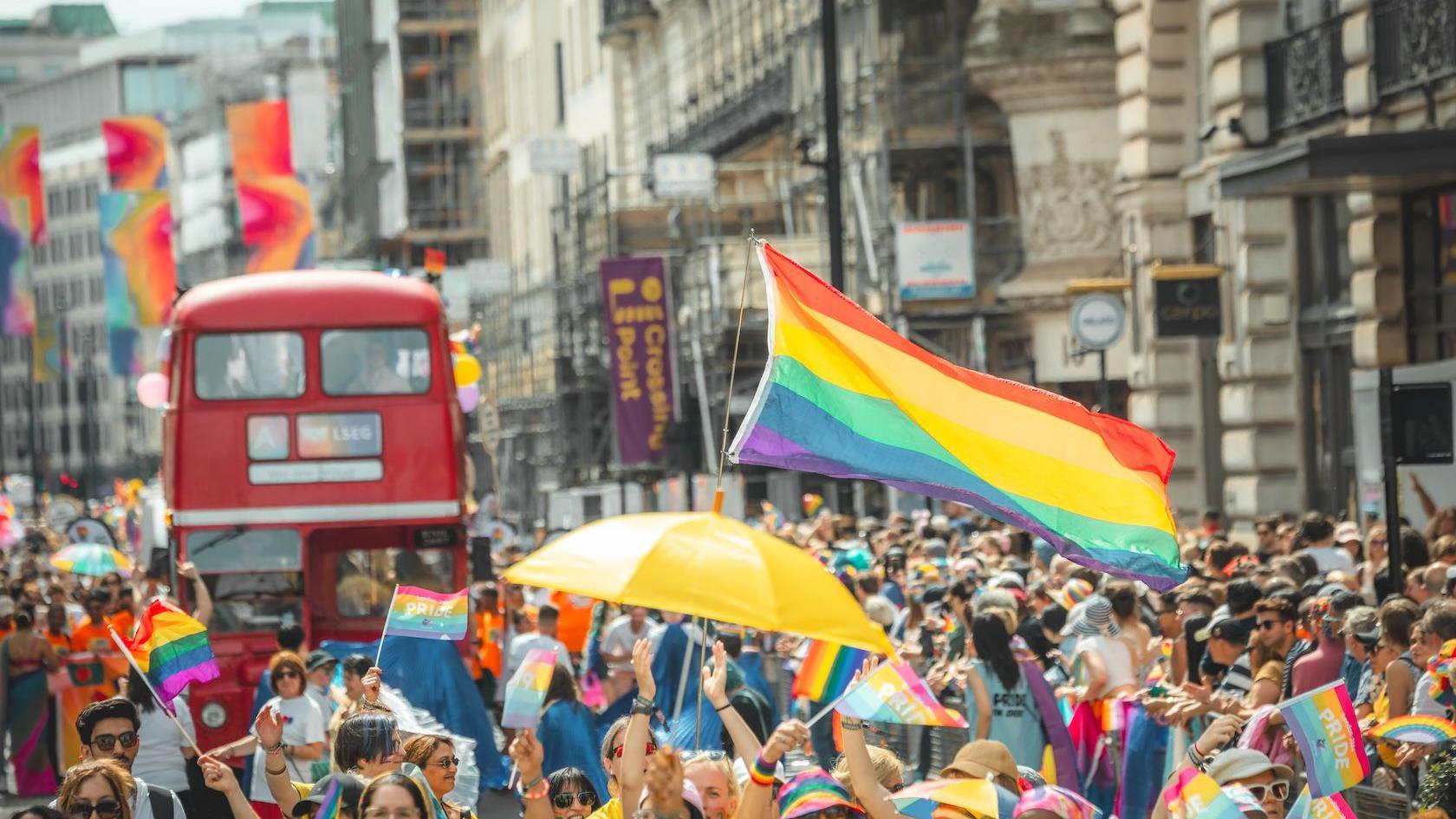 A Pride march with rainbow flags and a colourful umbrella fills a London street. A red bus adorned with colourful balloons follows behind the march while hundreds of spectators watch on. Rainbow flags hung from buildings can be seen in the background.