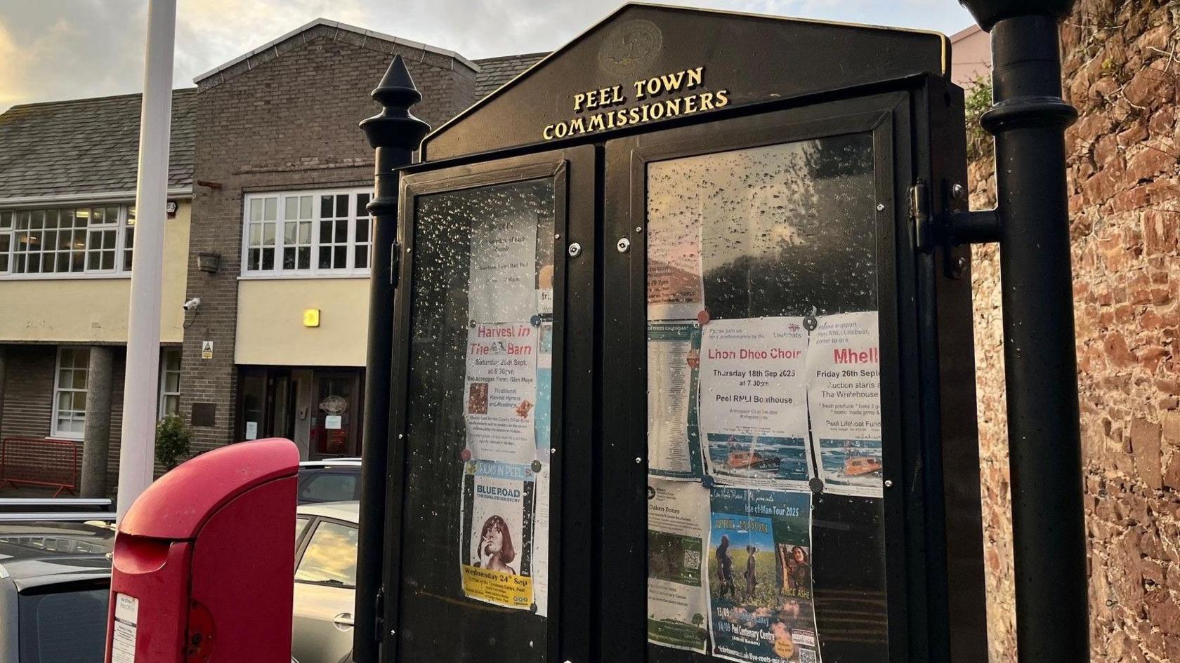 A black glass-fronted noticeboard on behalf of Peel Town Commissioners, featuring flyers and notices about community events. A red postbox stands to the left of the noticeboard and there are cars and buildings in the background.