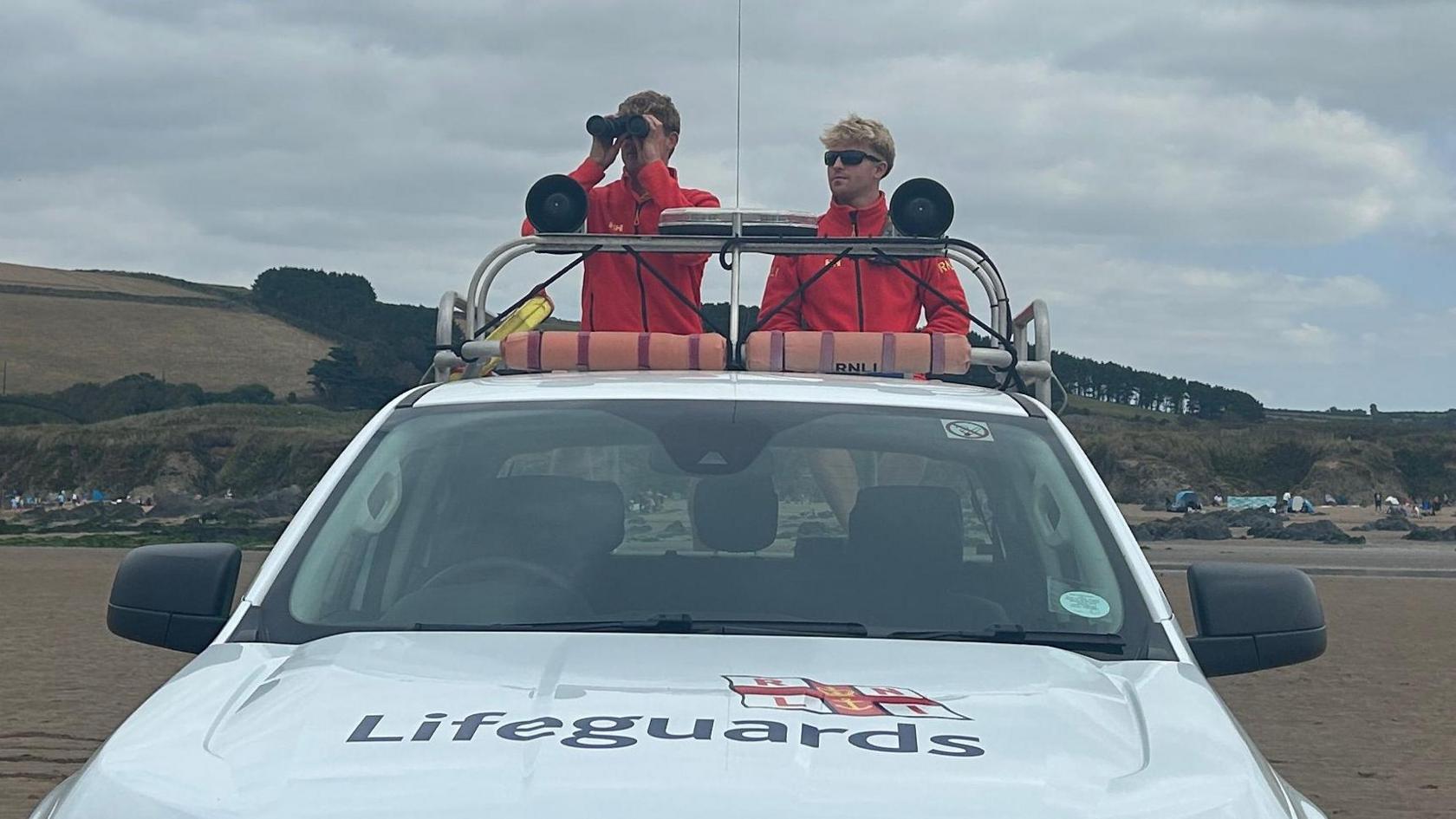 Two young male lifeguards in red jackets stand on the back of a white RNLI branded truck. They are on a sandy flat beach, with hills behind.