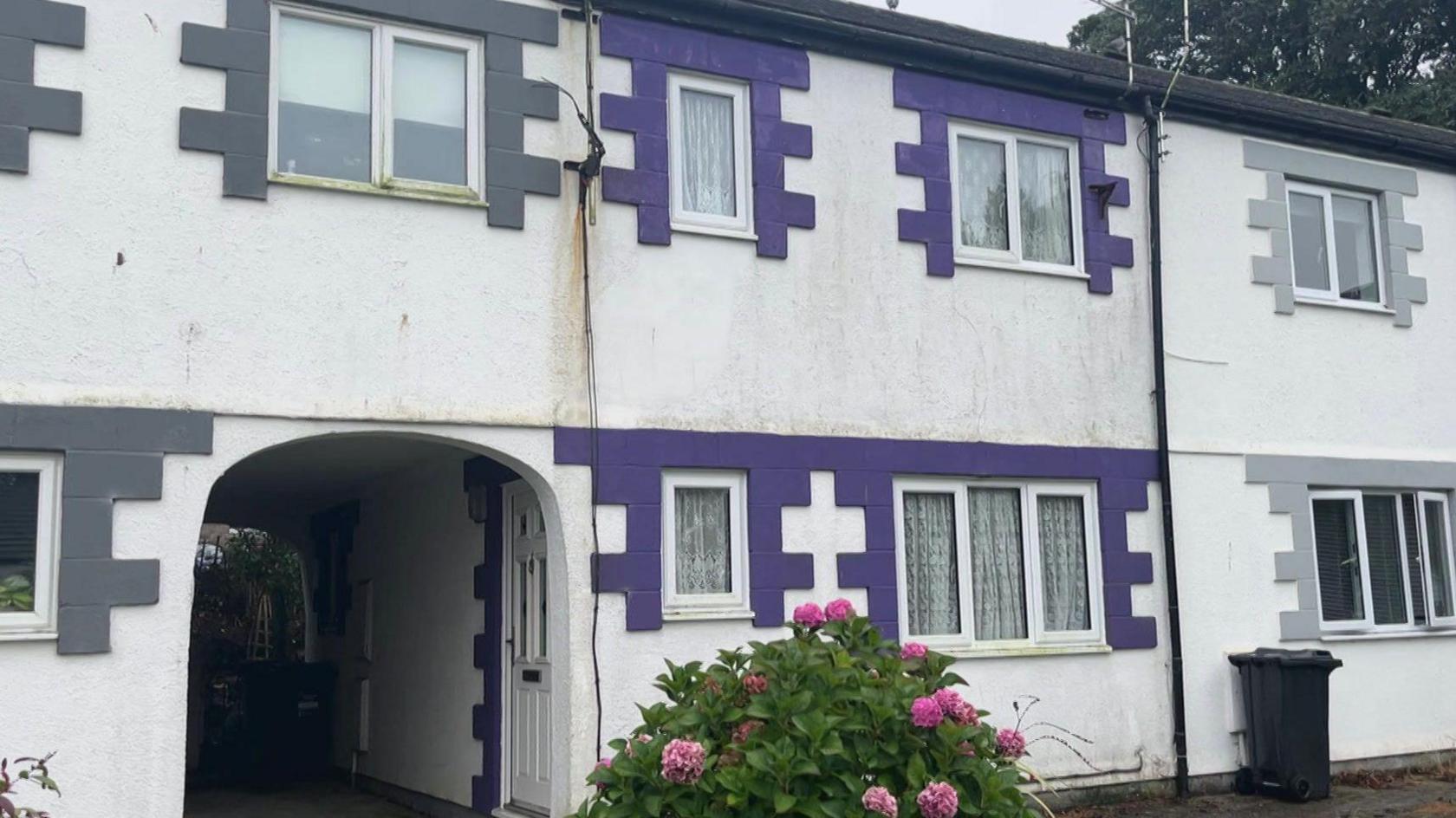 A small house in a courtyard in Hayle built in the 1980s. The door and windows have a distinctive purple trim. A pink hydrangea bush is outside.