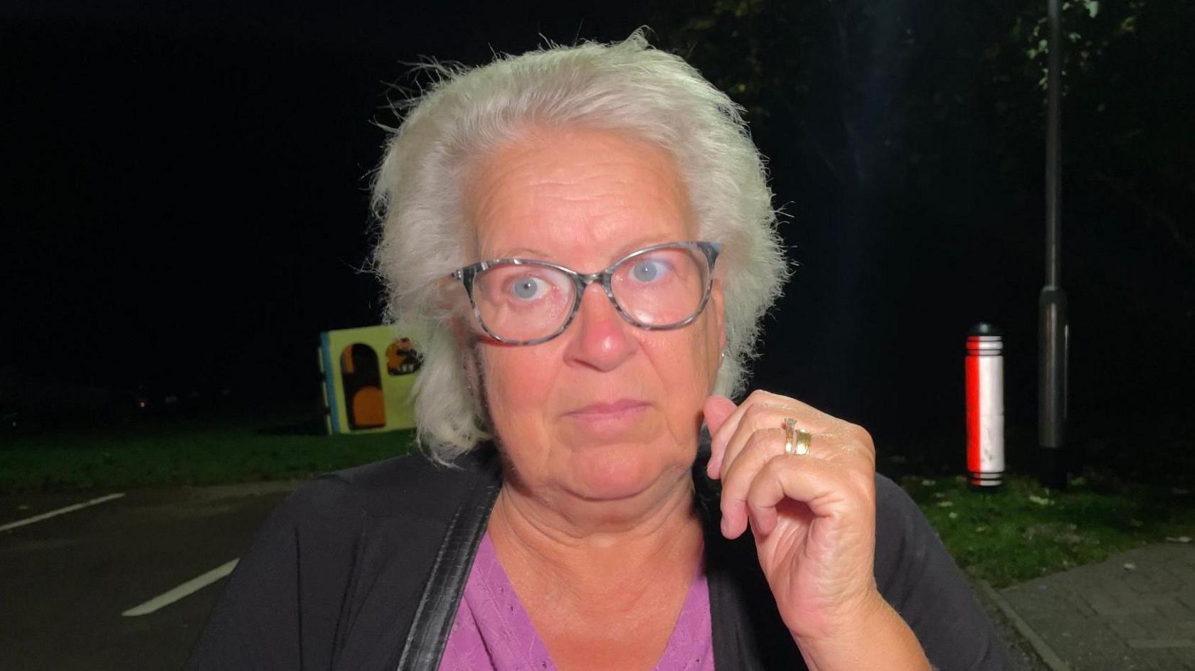 Susan Barnet pictured at night. She is wearing glasses and has a pink top on, and a black jacket. She has short, white hair. She is standing on a road. A verge and part of a streetlight can be seen on her right.