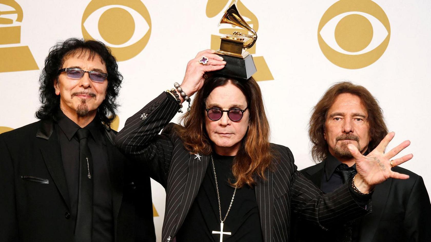 Bandmates Tony Iommi, Ozzy Osbourne and Geezer Butler pose in front of yellow and white Grammy logos while holding a grammy award for Best Metal Performance at the 2014 Grammy awards