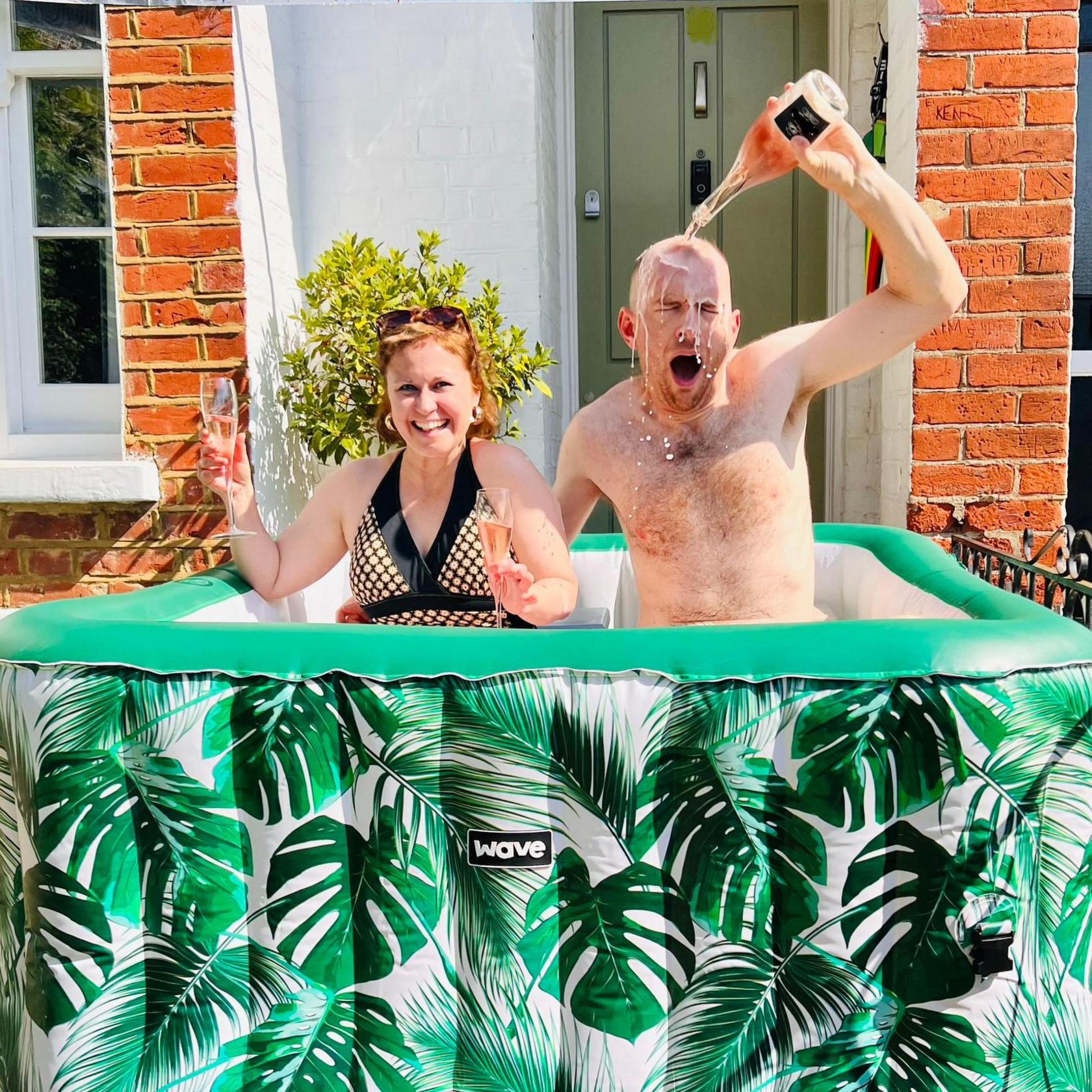 Tom and Beth sit in an inflatable palm-print inflatable pool. Beth is holding a glass of fizz in both hands and Tom is tipping the bottle of fizz over his head