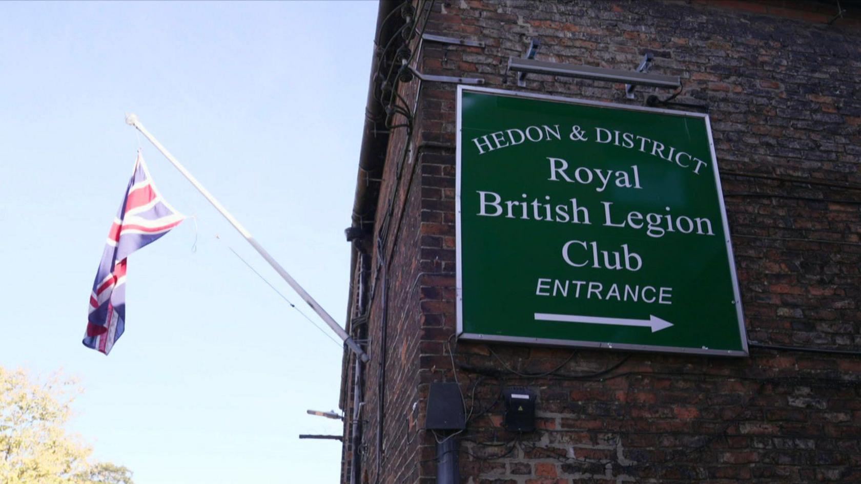 A square green sign reading 'Hedon & District Royal British Legion Club entrance' is on a brick wall. To the left of the sign is a Union Jack flag flying in the wind. On the sign there is an arrow pointing to the right.