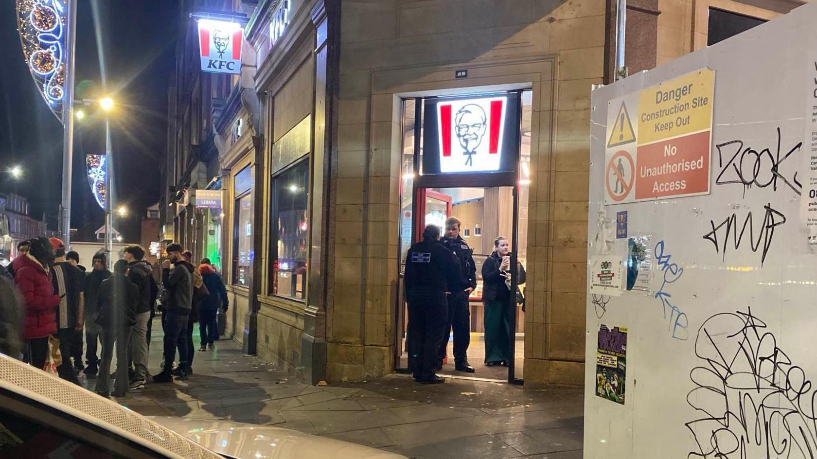 A parked police car near a KFC at night, with a number of officers stood in the doorway and members of the public loitering nearby on the pavement