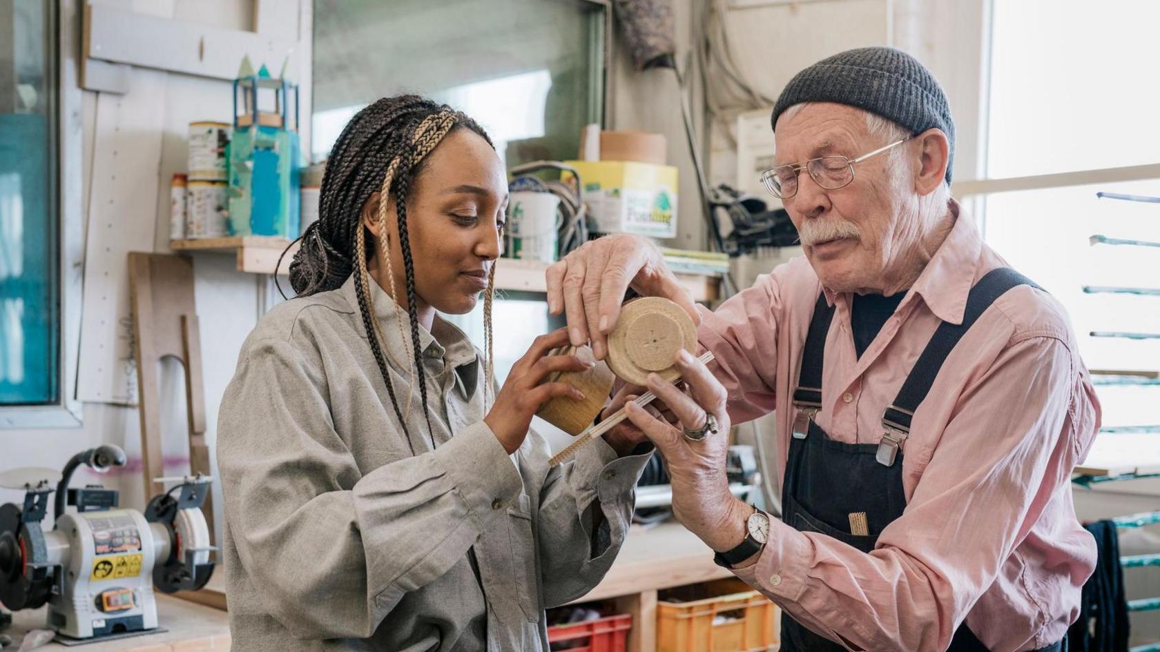 A young woman and an older man are both holding on to a wooden object. They are in a woodworking workshop with machinery and tools. The man is wearing a pink shirt and dungarees, the woman is wearing a grey shirt. 