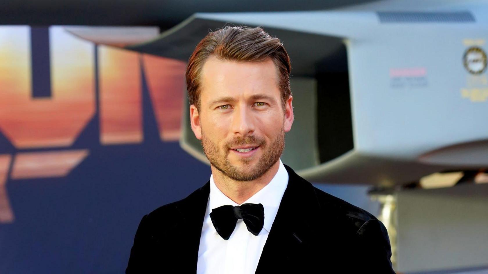 Glen Powell, dressed in a suit with bow tie, on the red carpet for a film premiere. He has dark brown hair and is unshaven with stubble. 