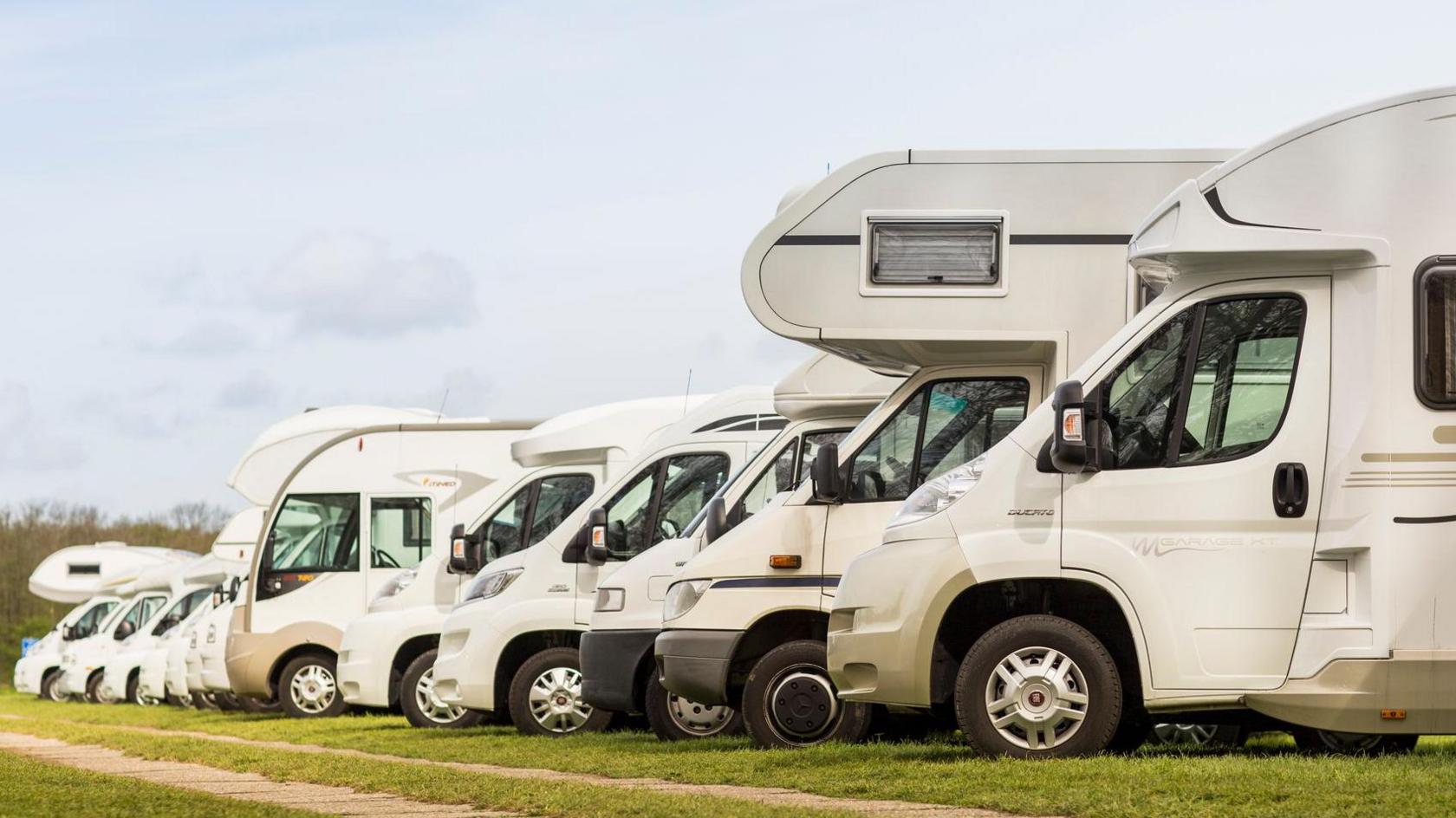 Side view of a row of motorhomes of varying size and design parked on a grass field with trees in the background