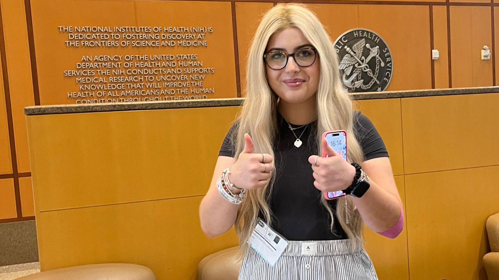 A woman with blonde hair stood in front of a brown wall and panels with signs and logos. She is holding two thumbs up to the camera, with her phone in one hand. The writing on the wall and accompanying test refers to The National Institutes of Health.