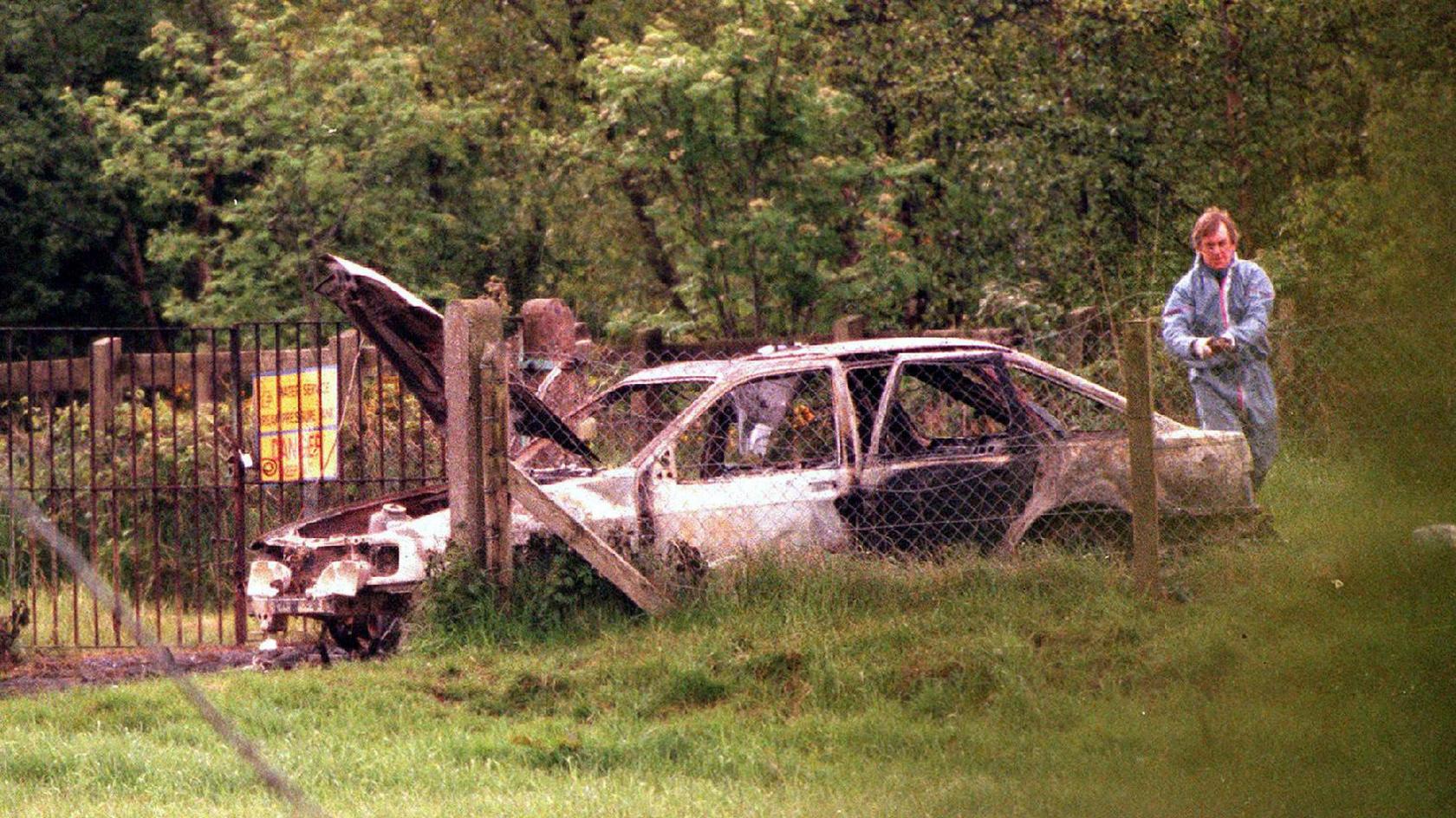 A burnt out car beside a wire fence. A forensic officer can be seen inspecting it. 