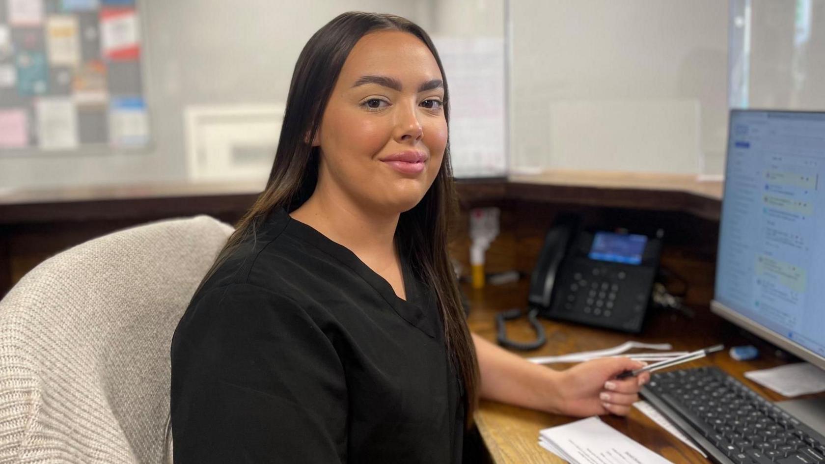 A receptionist in black scrubs sits at a desk in front of a computer