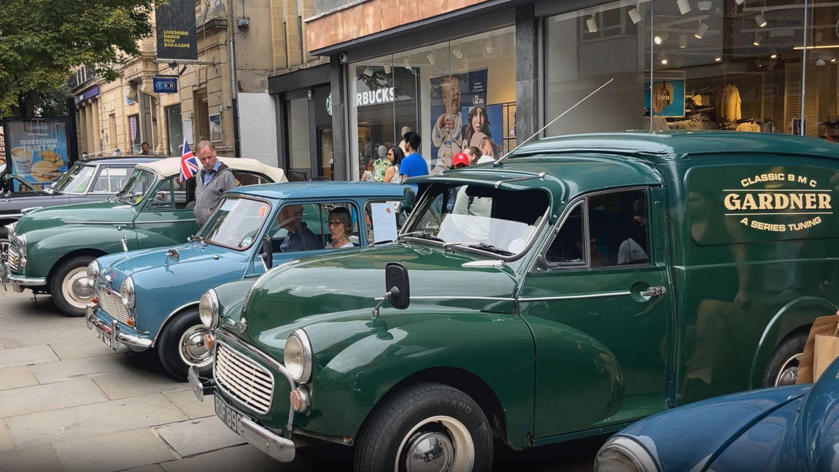 Classic cars and small vans in blue and green lined up along a street outside shops. They are faced outwards, on display.
