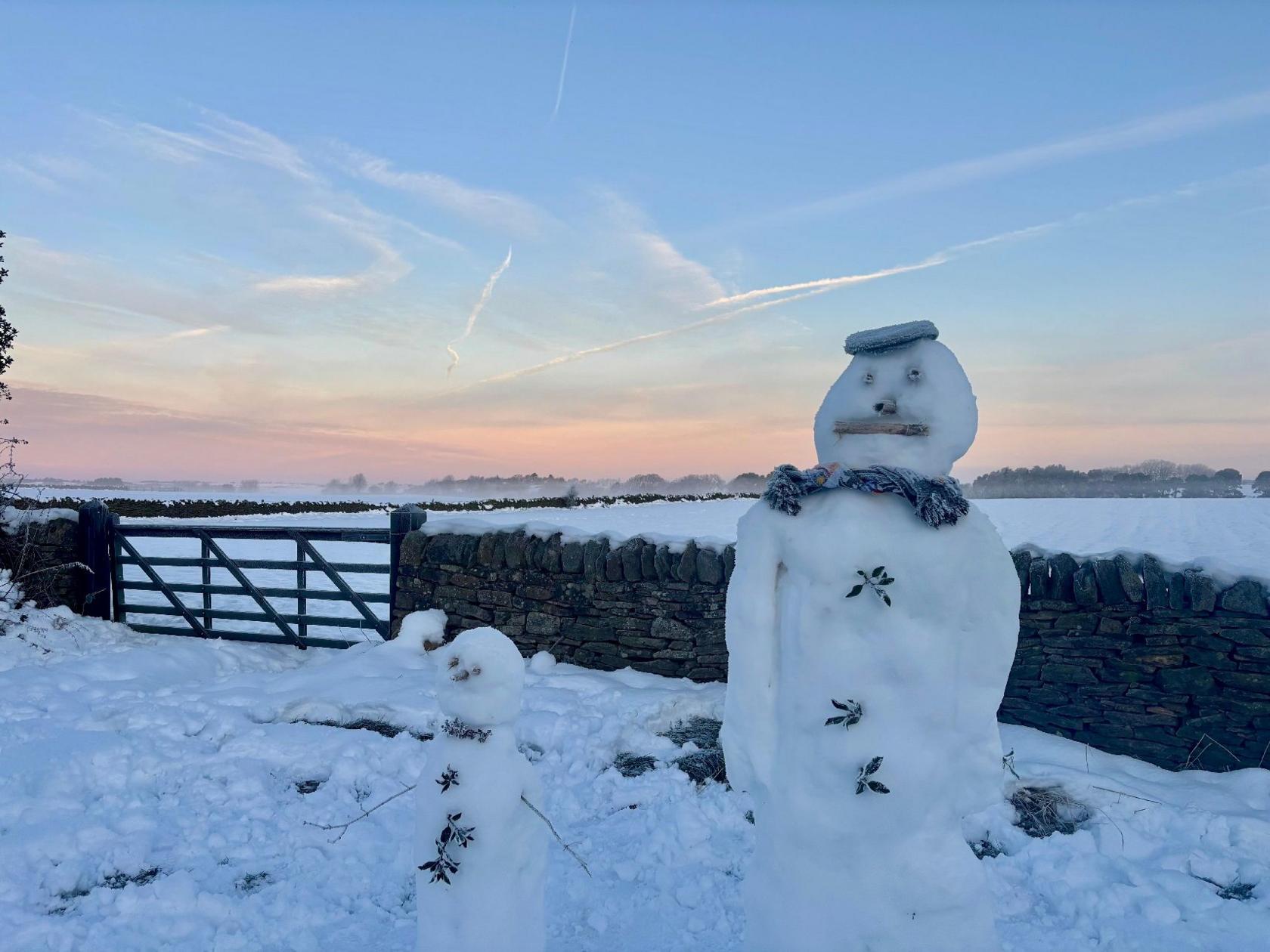 A small snowman beside a larger snowman in front of a stone wall and a field covered in snow