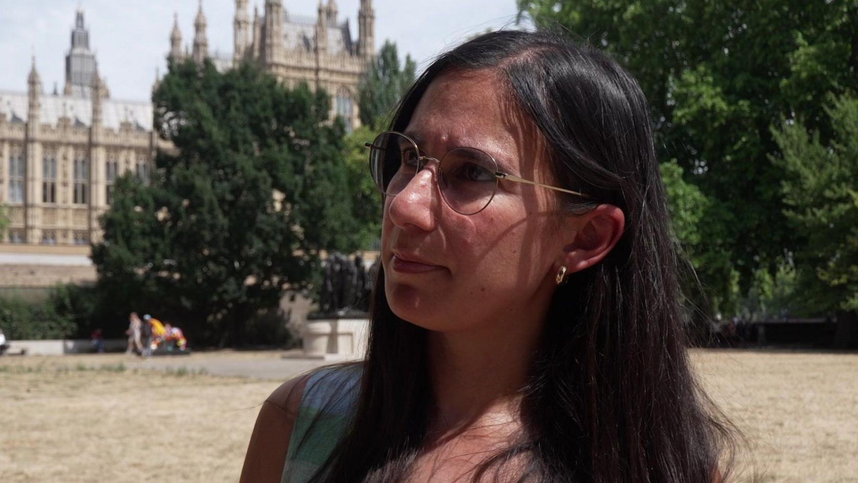 Ruth Fitzharris stands outdoors with the Palace of Westminster and trees visible behind her. Ruth Fitzharris has long dark hair and is wearing a sleeveless light-coloured top, while people can be seen in the distance.