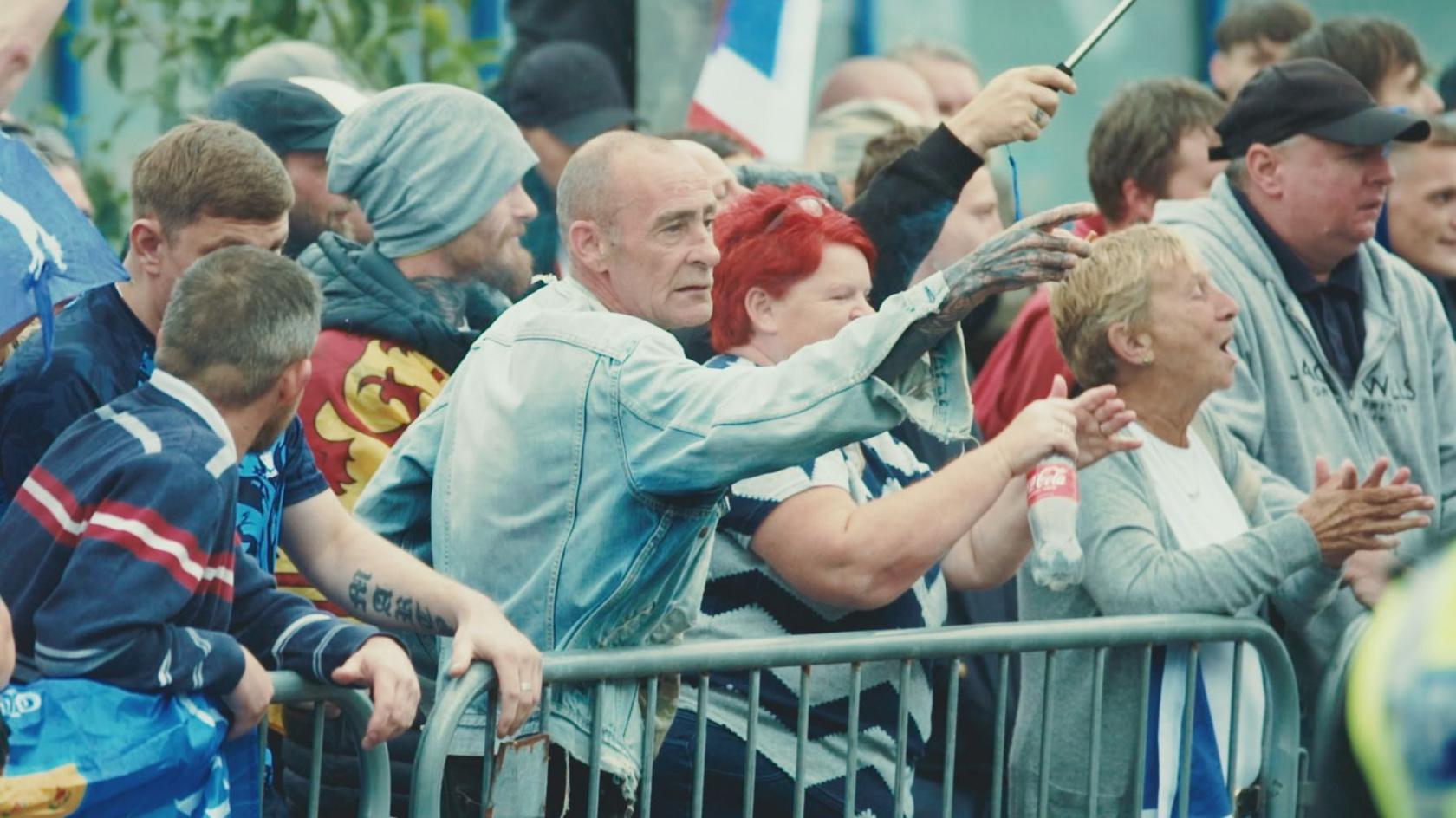 A number of protesters standing behind a barrier outside the hotel. One man in the middle, wearing a denim jacket, points with his right hand. Next to him two women are clapping.