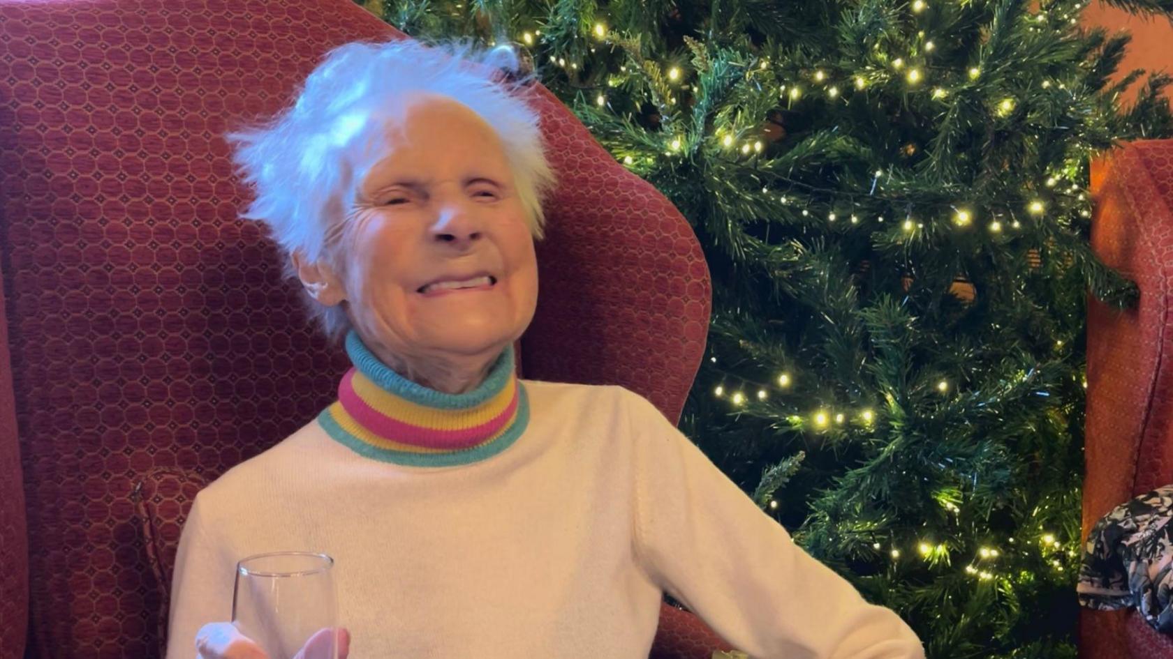 Molly, who is wearing a rainbow coloured jumper, is sitting in a red armchair and smiling. She is holding a champagne glass.