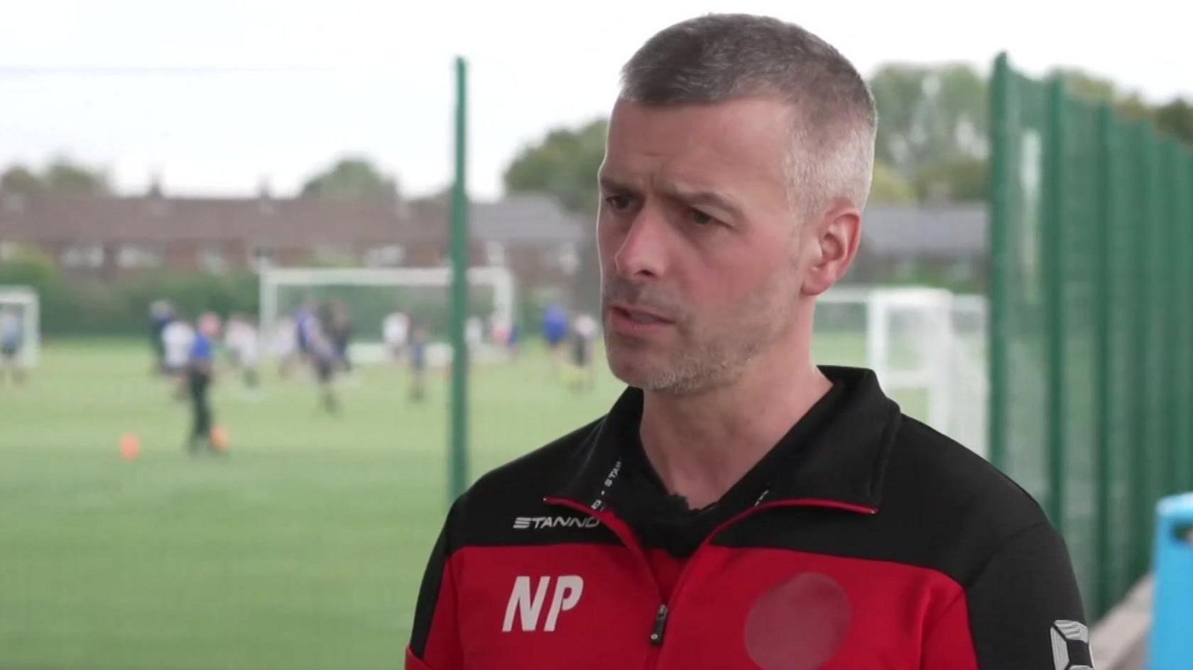 Neil Proctor has short hair. He is wearing a red and black t-shirt and a red and black tracksuit top.