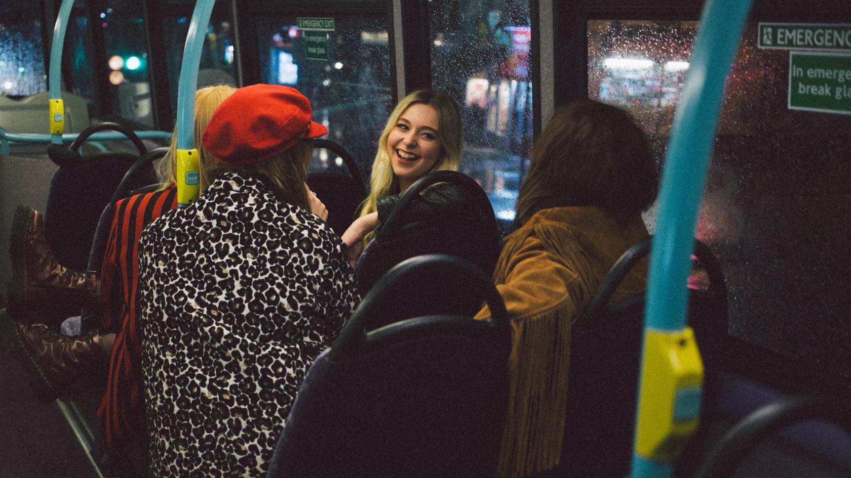 A stock image of some older teenage girls smiling and chatting on a bus, it is night time and it is raining outside. 