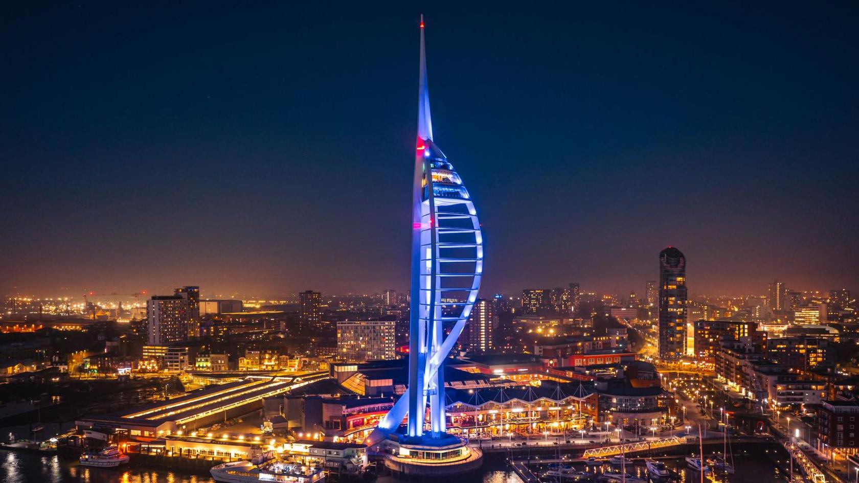 Aerial night view of the illuminated Spinnaker Tower in Portsmouth, glowing in blue and red lights with reflections shimmering on the water and surrounding city buildings lit up in the background.